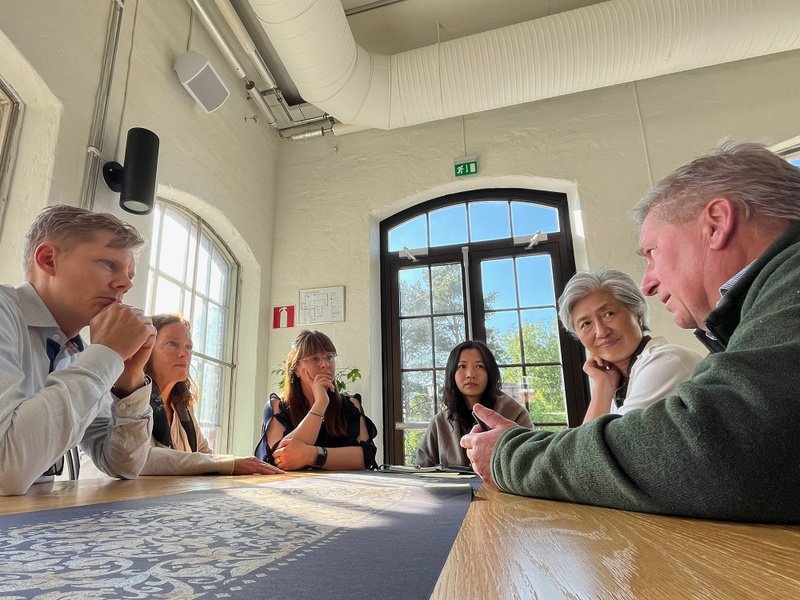 Sex personer sitter runt ett bord i ett ljust rum med stora fönster och diskuterar. En man i grön fleece talar medan de andra lyssnar uppmärksamt. Ljuset från fönstret lyser upp ansiktena.