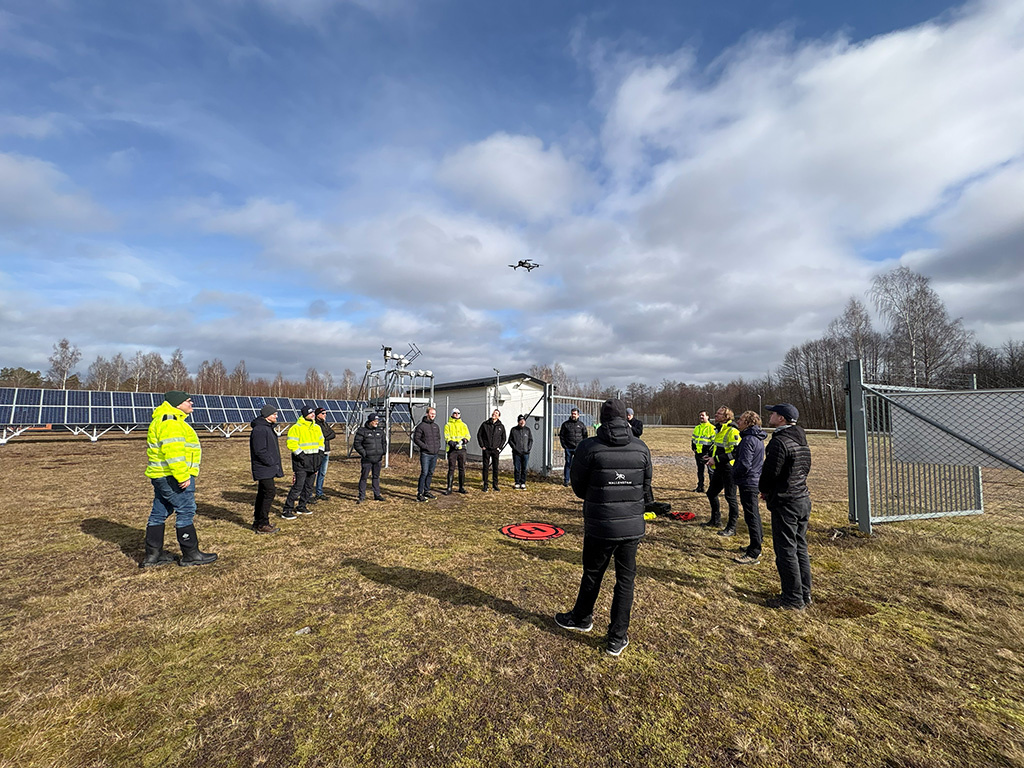 Personer står i en solpark och tittar på en drönare som lyfter från en röd startplatta.