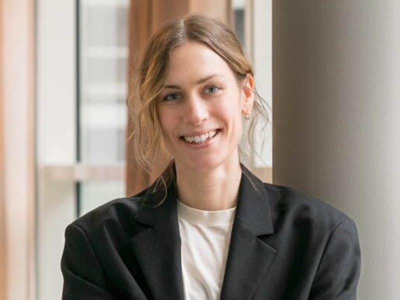 Porträtt av en ung kvinna i vit tröja och svart kavaj som står framför ett fönster.