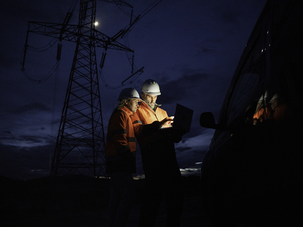 Two persons standing with a laptop in front of a power grid.