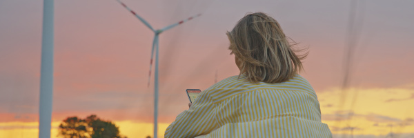 A woman sitting on the grass, looking at her mobile phone, with wind turbines on the horizon.