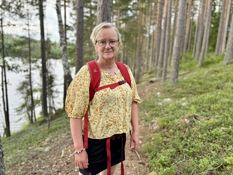 En kvinna med glasögon, gulblommig blus, svart kjol och röd ryggsäck står på en skogsstig i tallskog med en sjö i bakgrunden.