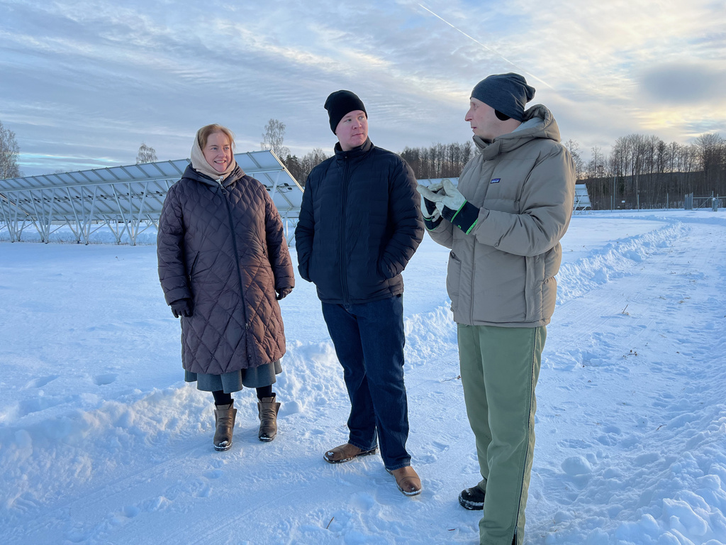 One women and two men stand outside in the snow in front of rows of solar energy panels.