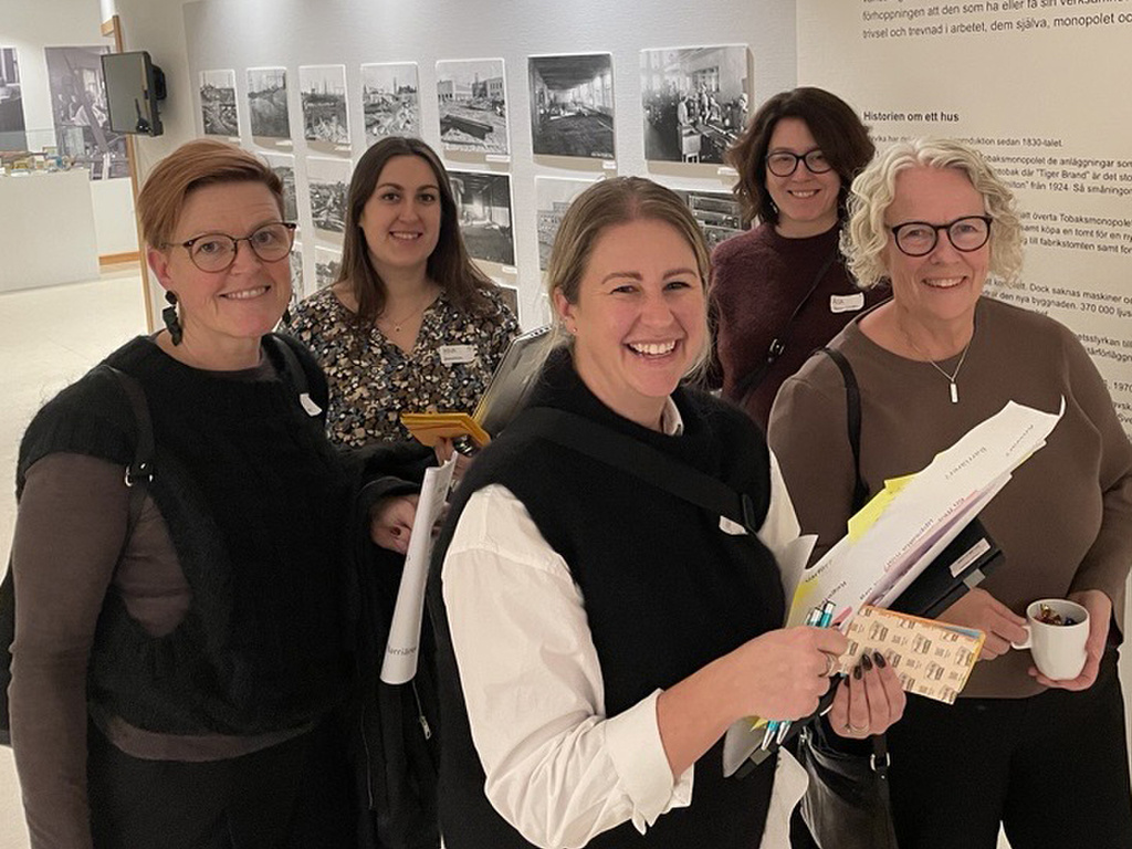 Fem kvinnor står i en ljus korridor framför små svartvita tavlor.