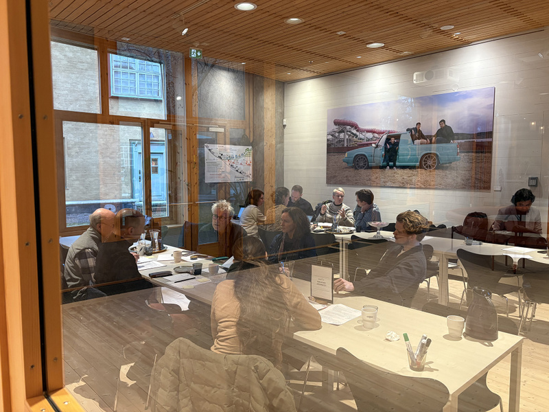 Människor sitter och diskuterar vid två långbord i ett rum med stora fönster. 