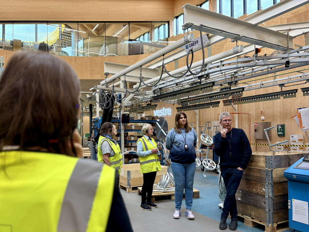 Personer i gula västar lyssnar till en man och kvinna som talar i en fabrik.