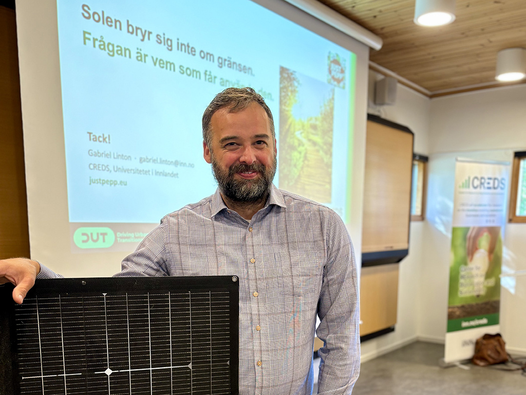 En man med skägg står och ler med handen på en solpanel framför en stor digital tavla.