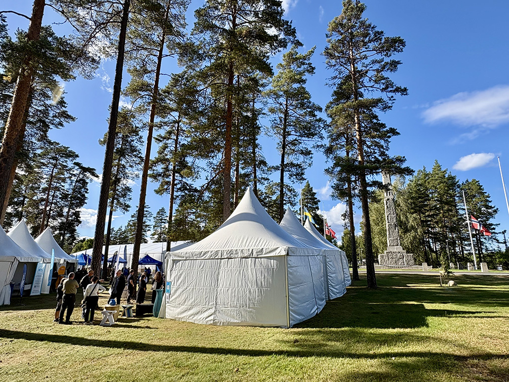 Vita tält på en grön gräsmatta bland höga tallar mot en klarblå himmel. I bakgrunden syns svenska och norska flaggor och ett högt monument i sten.