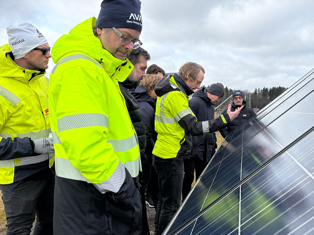 Kursdeltagare i gula varseljackor och mössor tittar närmare på en solpanel i en solcellsanläggning utomhus.