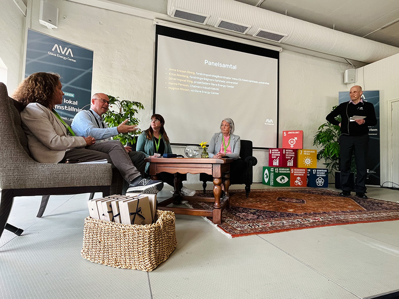 Fem personer deltar i ett panelsamtal i en ljus lokal. Fyra personer sitter i fåtöljer kring ett soffbord medan en femte person står och håller i ett papper. I bakgrunden syns en presentationsskärm med namnen på deltagarna och en stapel med färgglada kuber som symboliserar globala hållbarhetsmål.