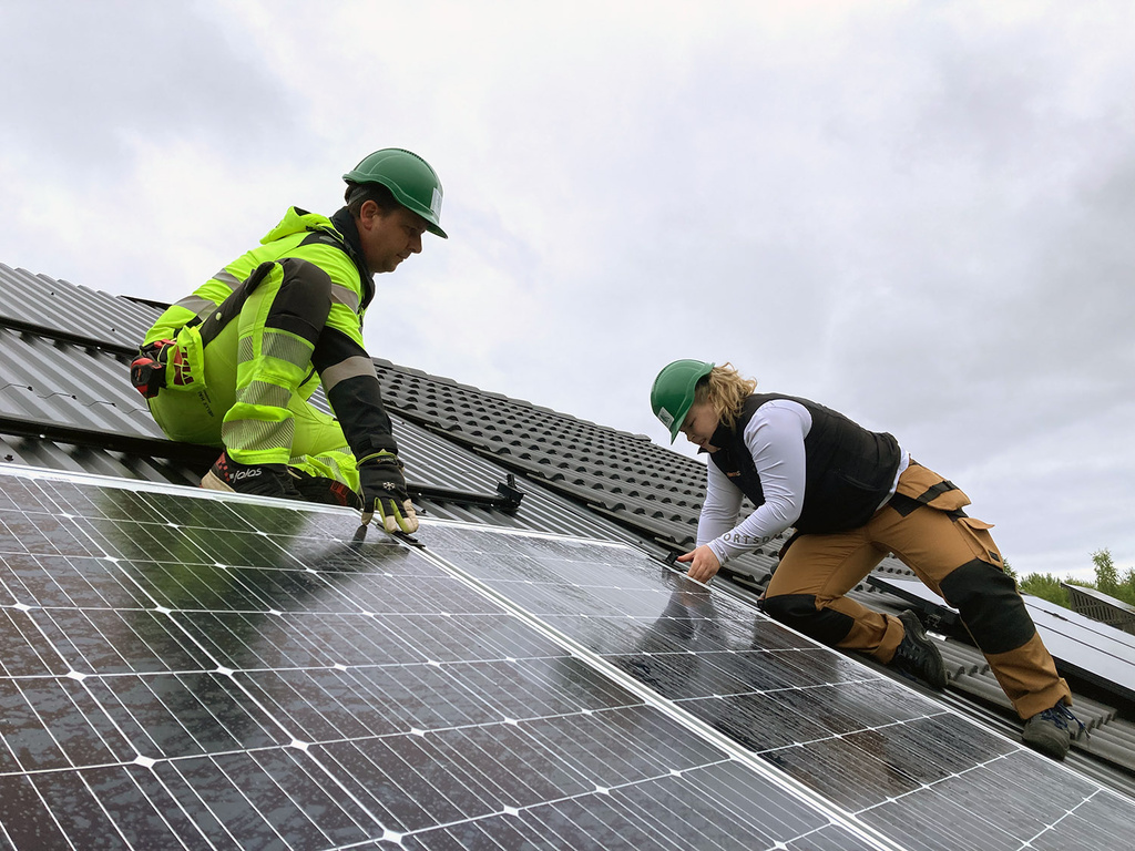 A man and a woman in work clothes and green helmets are sitting on a roof, inspecting the installation of a solar panel.