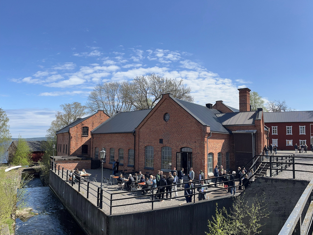 En grupp människor samlade utanför en röd tegelbyggnad vid en forsk och en sjö under blå himmel.