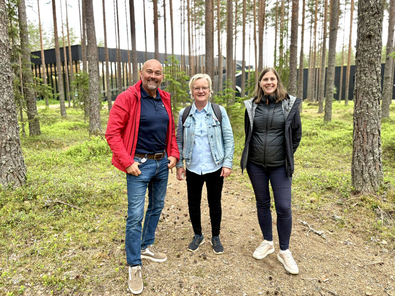 Tre personer står i en tallskog utanför en svart byggnad i trä.
