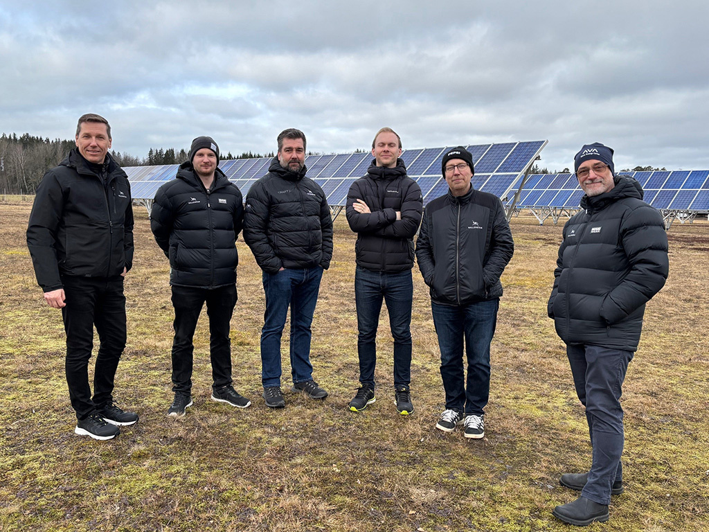 Sex män i svarta dunjackor står bredvid varandra och tittar in i kameran utomhus i en solpark.