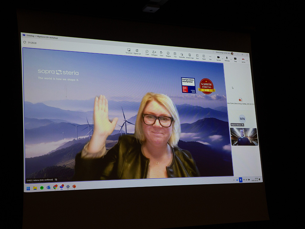 En kvinna med ljust hår och glasögon syns på en datorskärm och hälsar genom att lyfta handen.