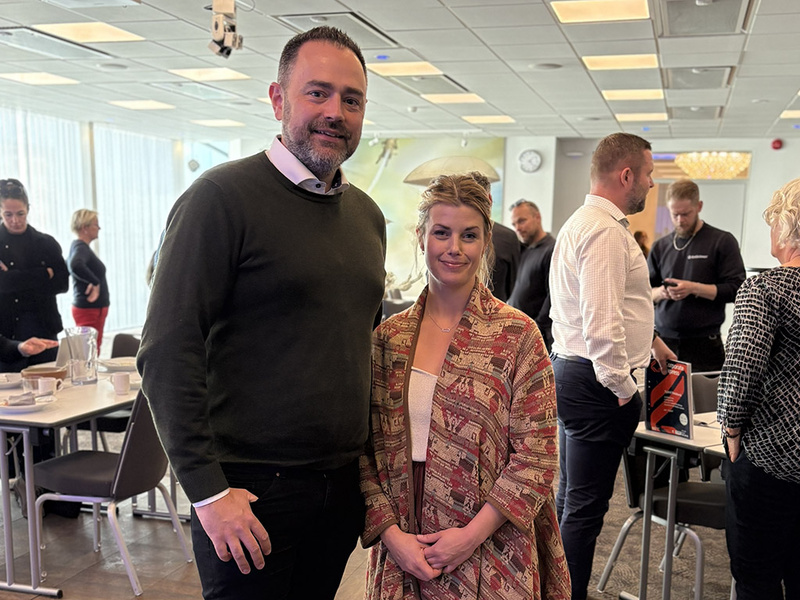 En man och en kvinna står i ett konferensrum med andra människor i bakgrunden.