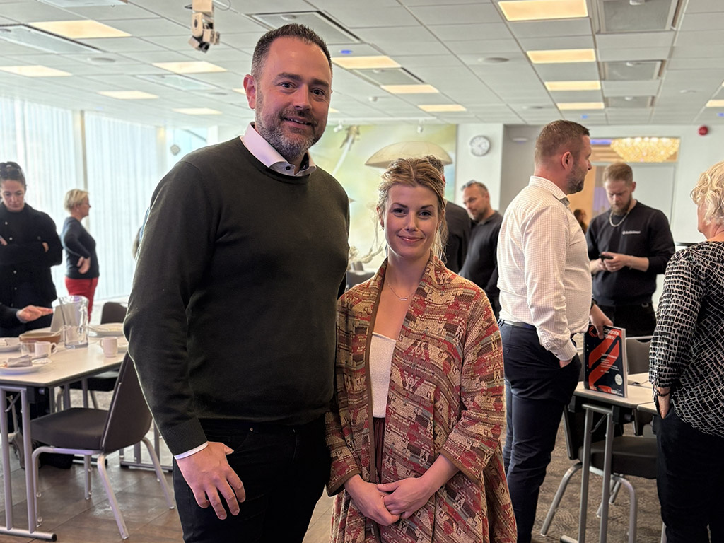 En man och en kvinna står i ett konferensrum med andra människor i bakgrunden.